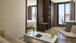 A modern hotel room with a bed, a dining area, a small kitchen, and a balcony with a city view.