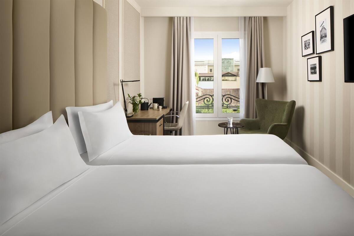 A hotel room with two beds, a desk, a chair, a lamp, and a balcony with a view.