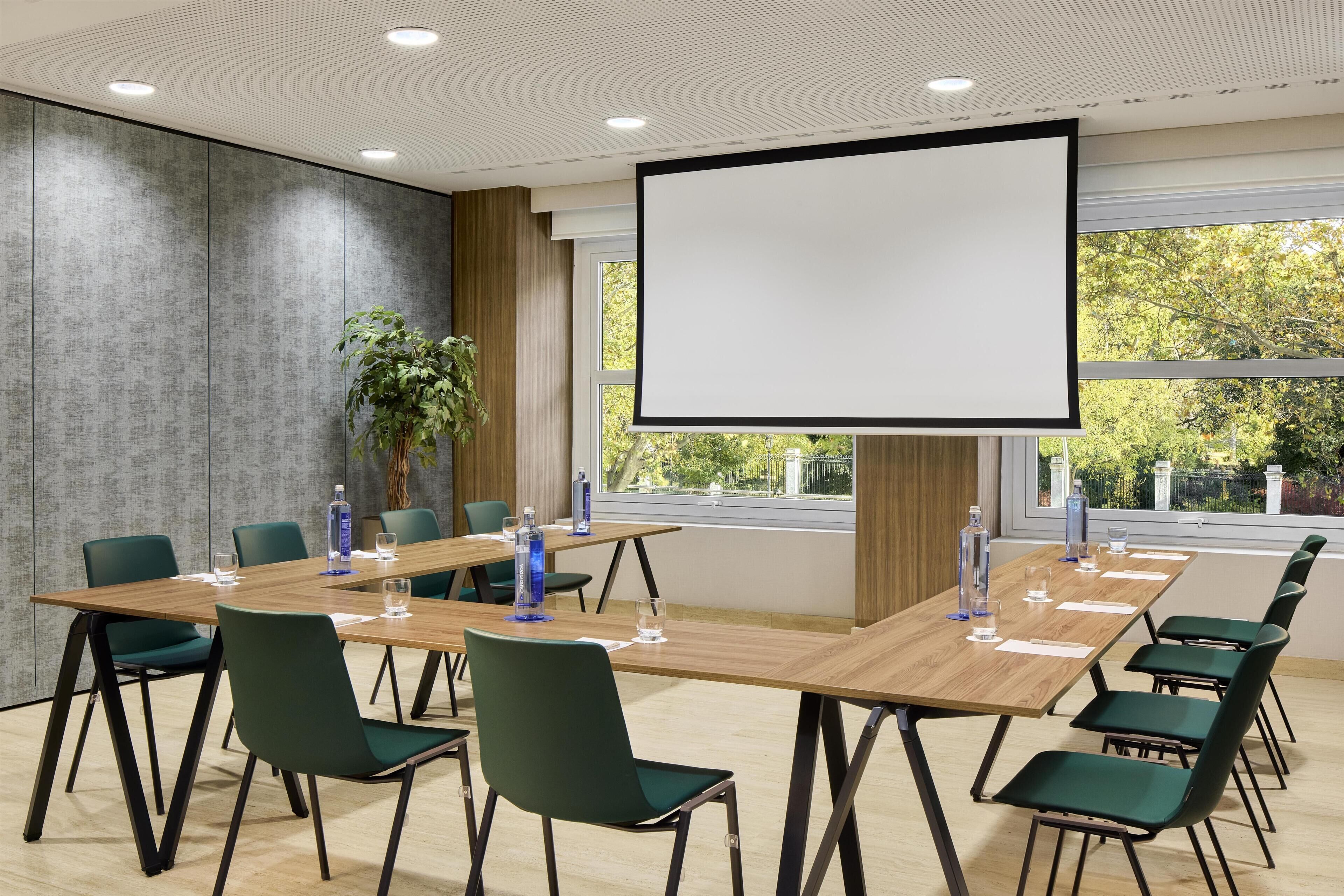 A conference room with a large screen, green chairs, a wooden table, and a potted plant.