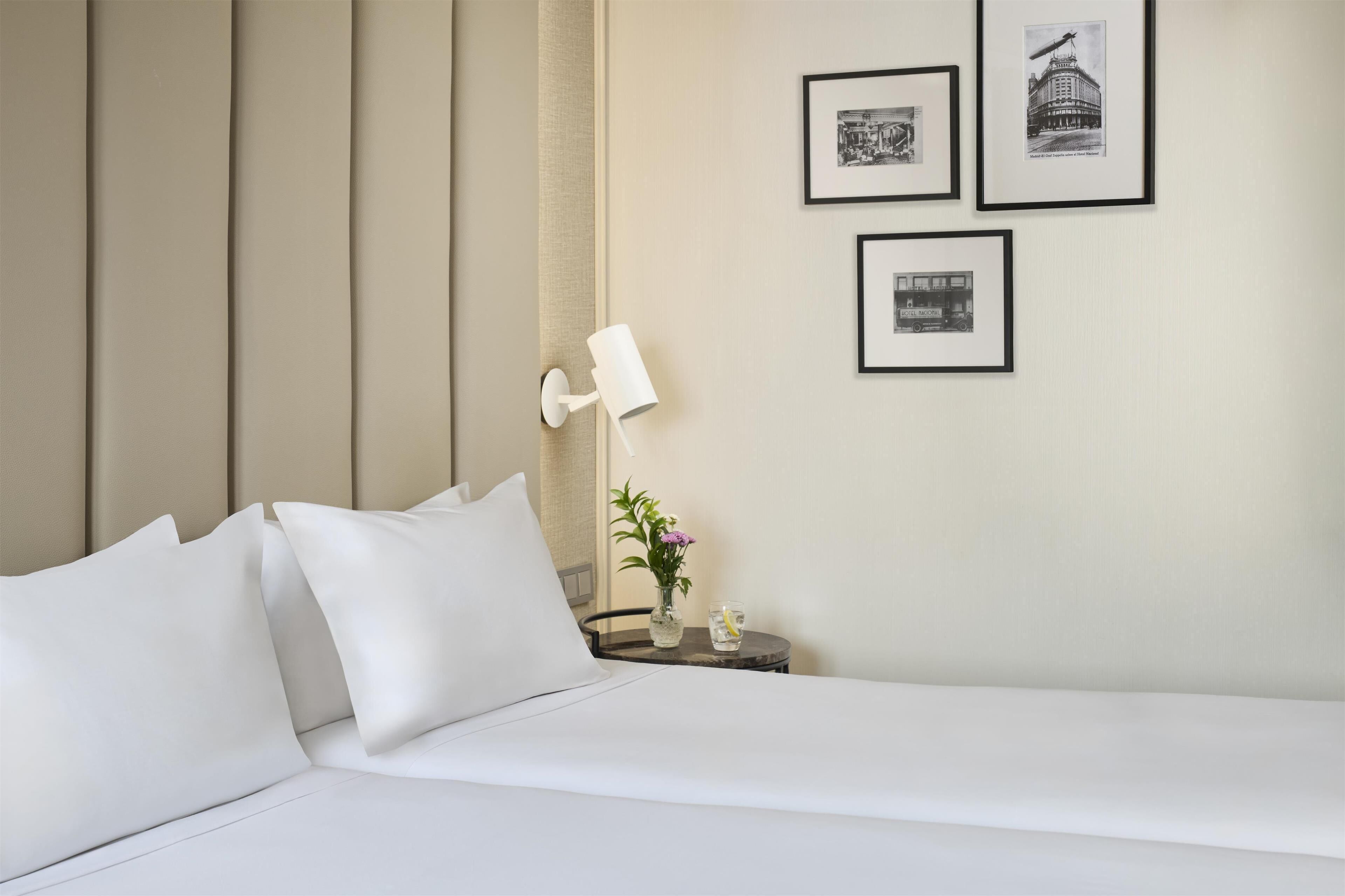 Una cama bien hecha con sábanas y almohadas blancas, una mesita de noche con una lámpara y un florero con flores, y tres cuadros en la pared.