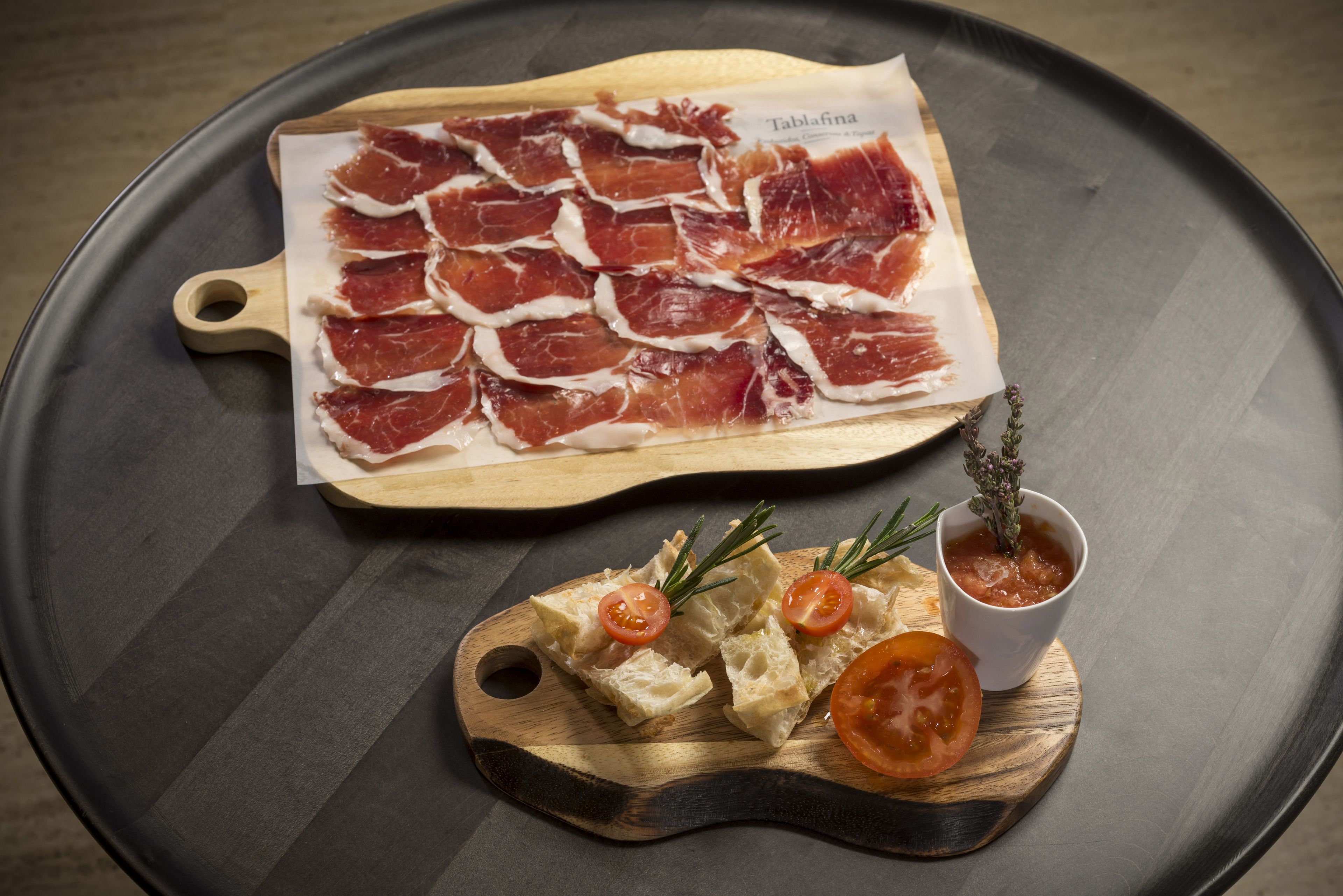 A wooden cutting board with prosciutto and a small bowl of sauce, placed on a round table.