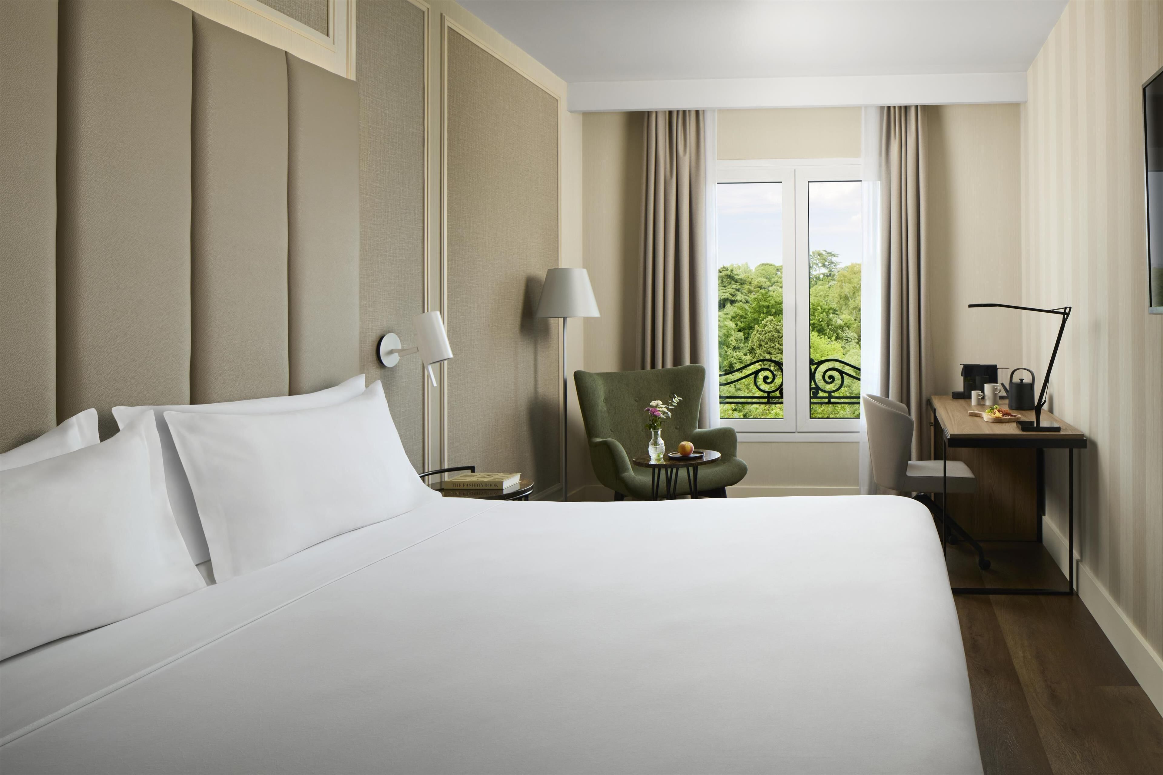 A hotel room with a large bed, a desk, a chair, and a view of greenery outside.