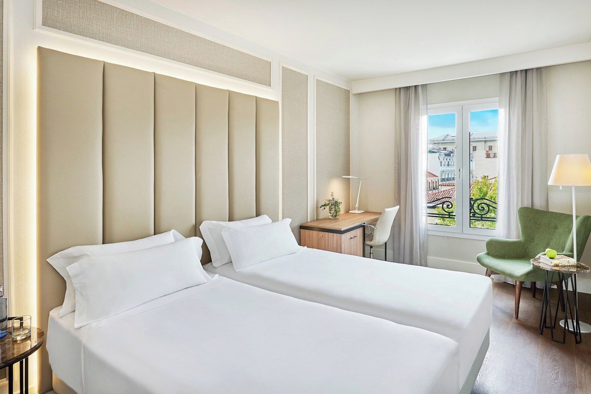 A hotel room with two beds, a desk, a chair, a lamp, and a view of buildings.
