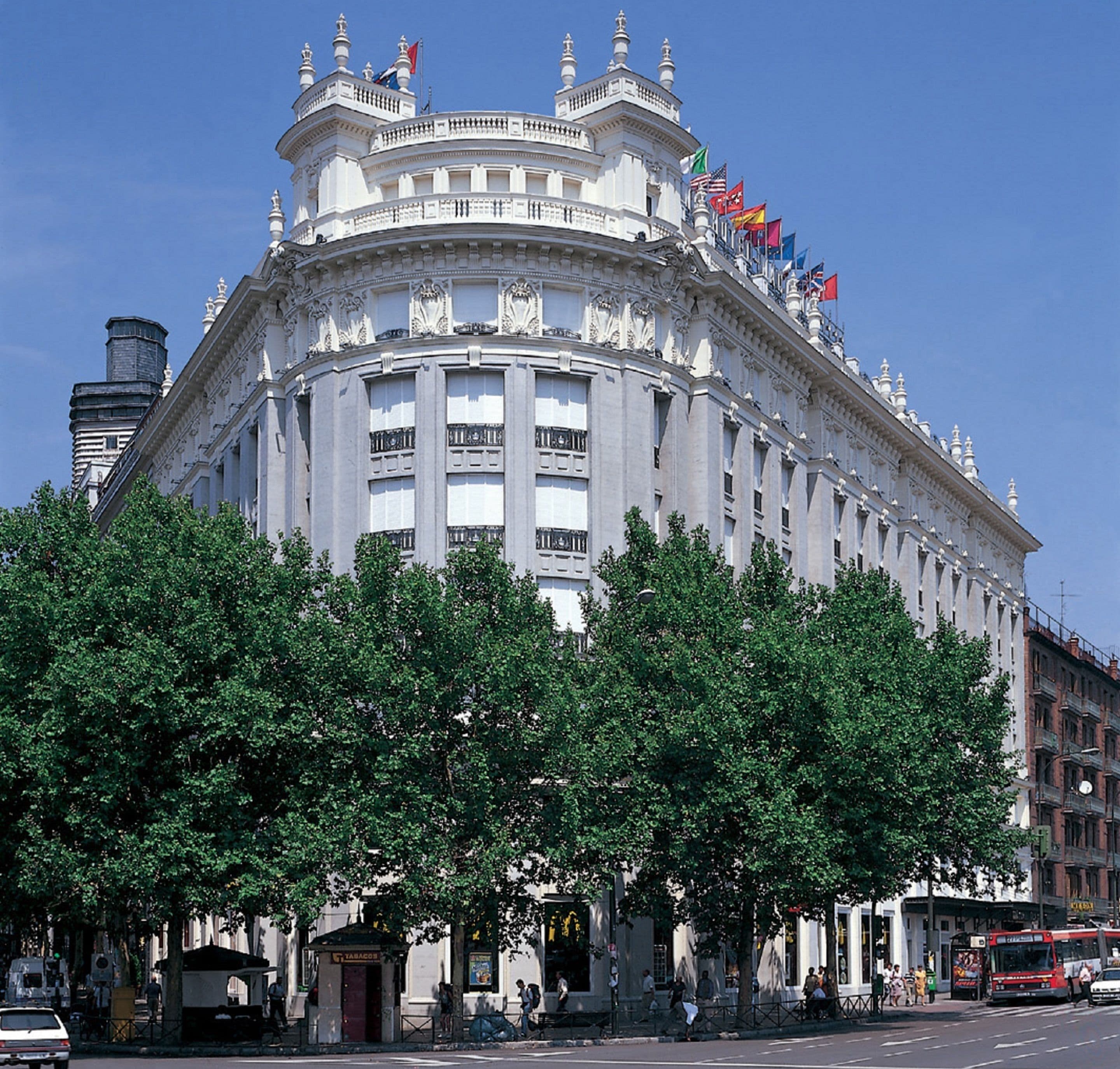 A large, ornate building with multiple flags, surrounded by trees and a street scene.