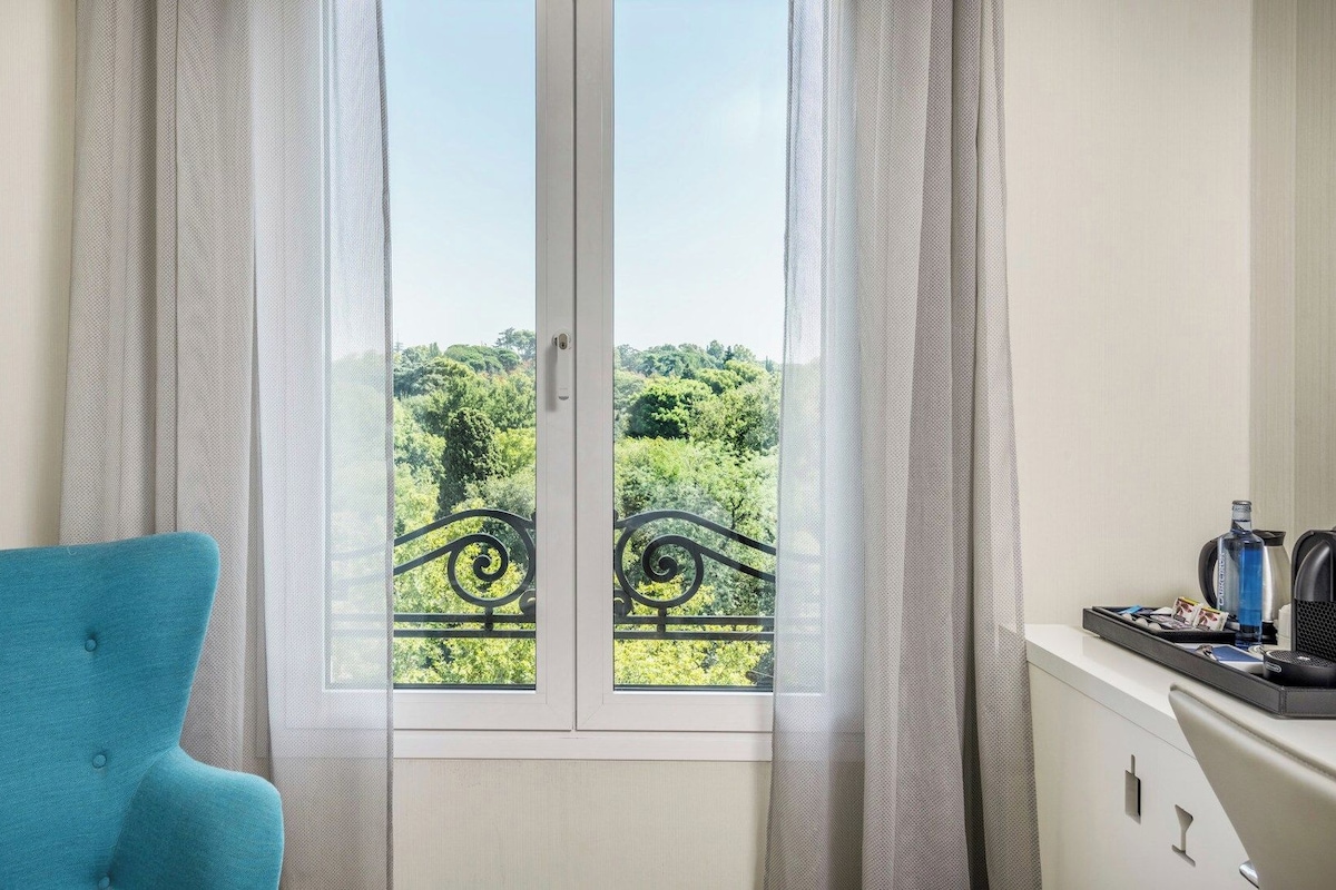 A room with a view of greenery through a window with a metal railing.