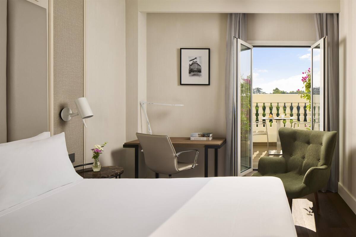 A hotel room with a bed, desk, chair, and a balcony with a view.