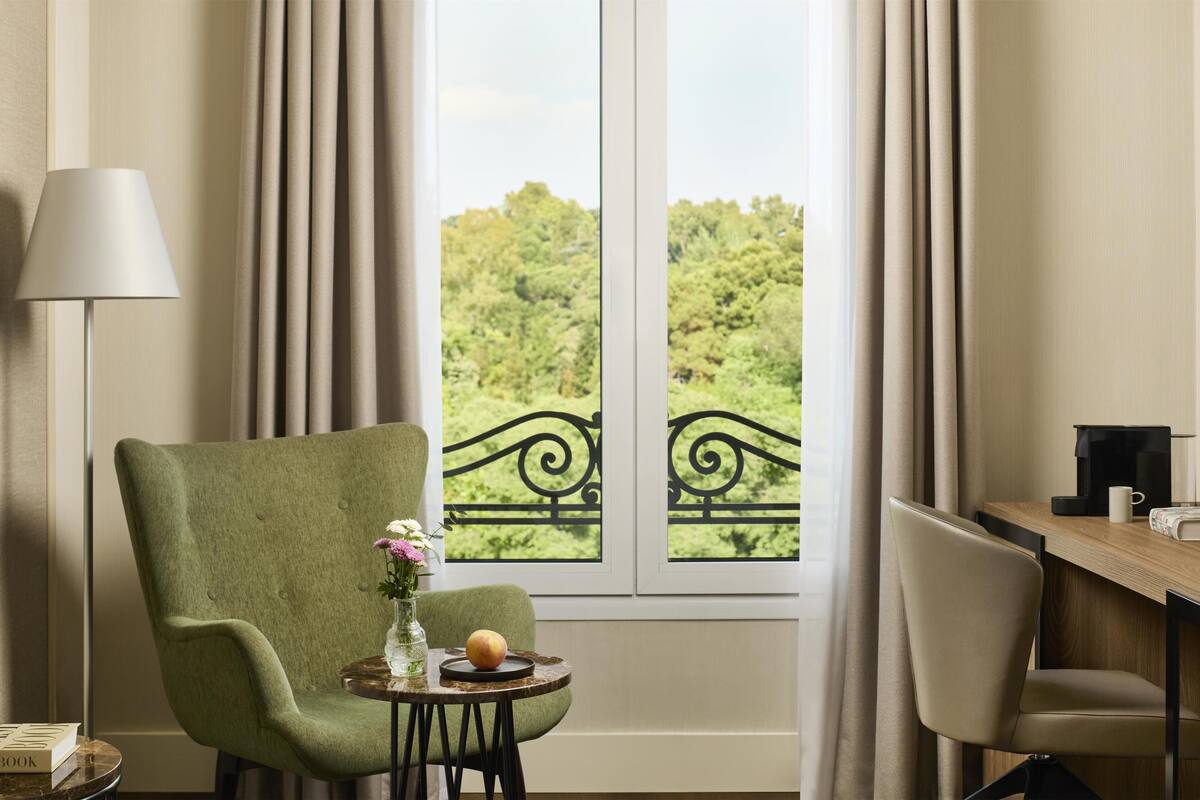 A room with a green armchair, a small round table with a vase and fruit, and a view of greenery through a window with curtains.