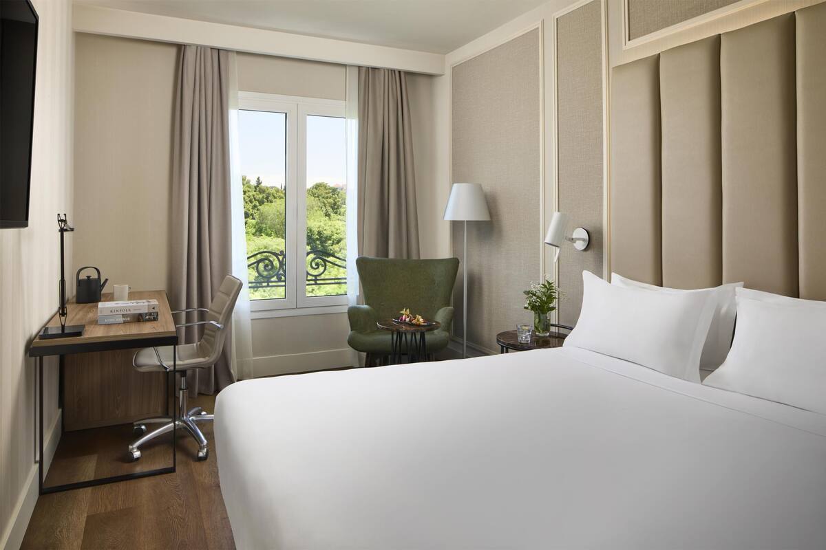A hotel room with a bed, a desk with a chair, a lamp, and a view of greenery outside.