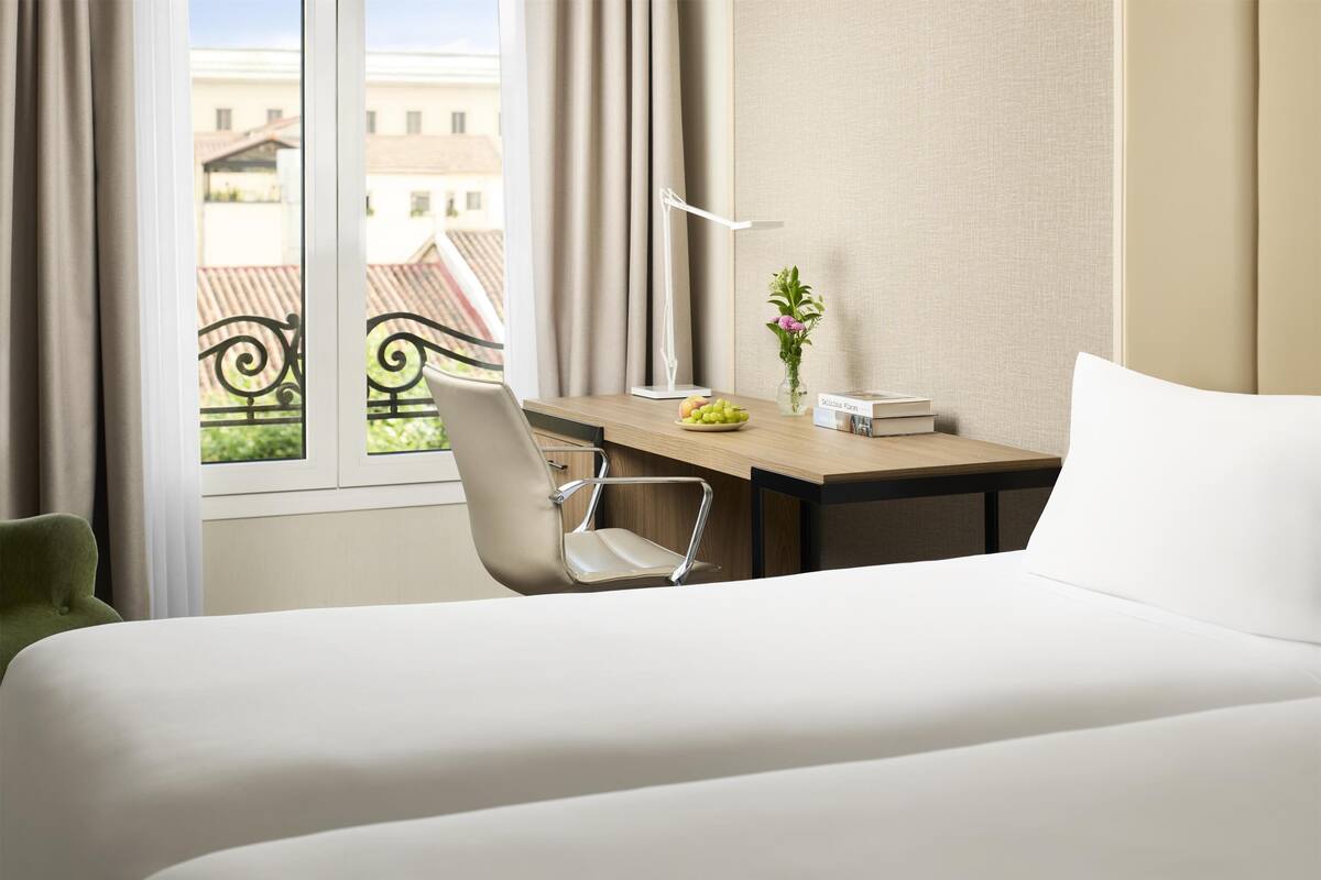A hotel room with a bed, a desk with a chair, a vase with flowers, and a view of buildings through the window.