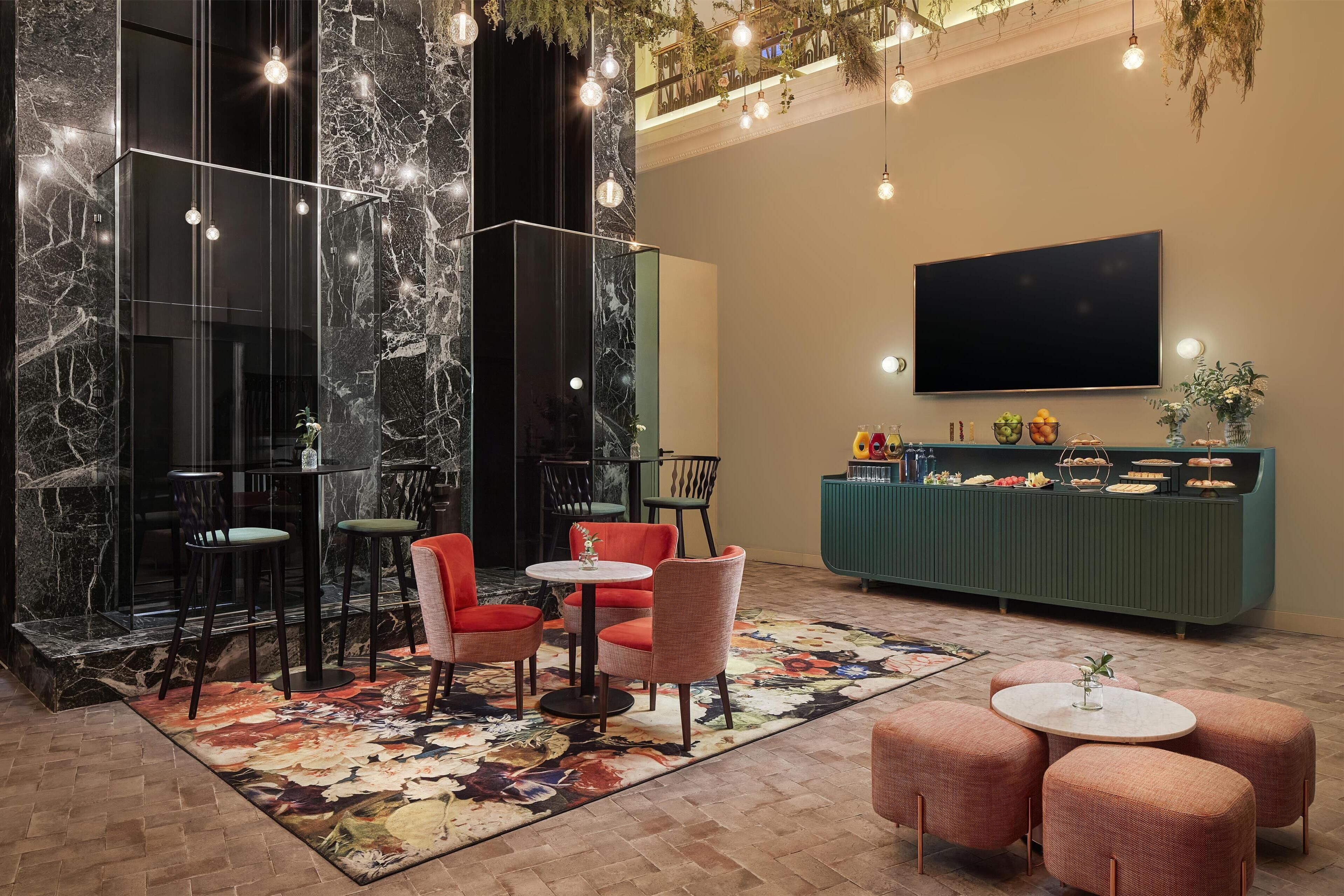A modern lobby with marble walls, a green counter with food and drinks, and seating areas with round tables and chairs.