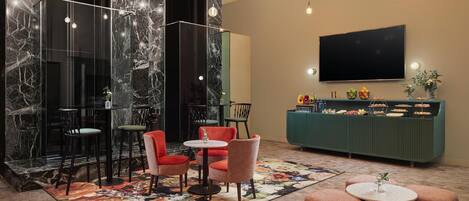 A modern lobby with marble walls, a green counter with food and drinks, and seating areas with round tables and chairs.