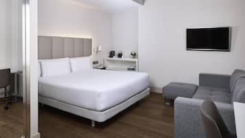 Una habitación de hotel moderna con una cama grande, un sofá gris, un televisor de pantalla plana montado en la pared y una mesita de noche con una lámpara.