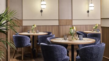 Un comedor de hotel con mesas redondas, sillas tapizadas en azul y luminarias fijadas a la pared.