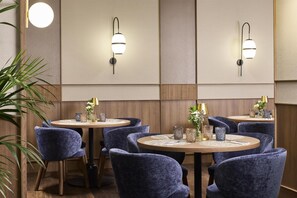 A hotel dining area with round tables, blue upholstered chairs, and wall-mounted light fixtures. - NH Sants Barcelona (Barcelona)