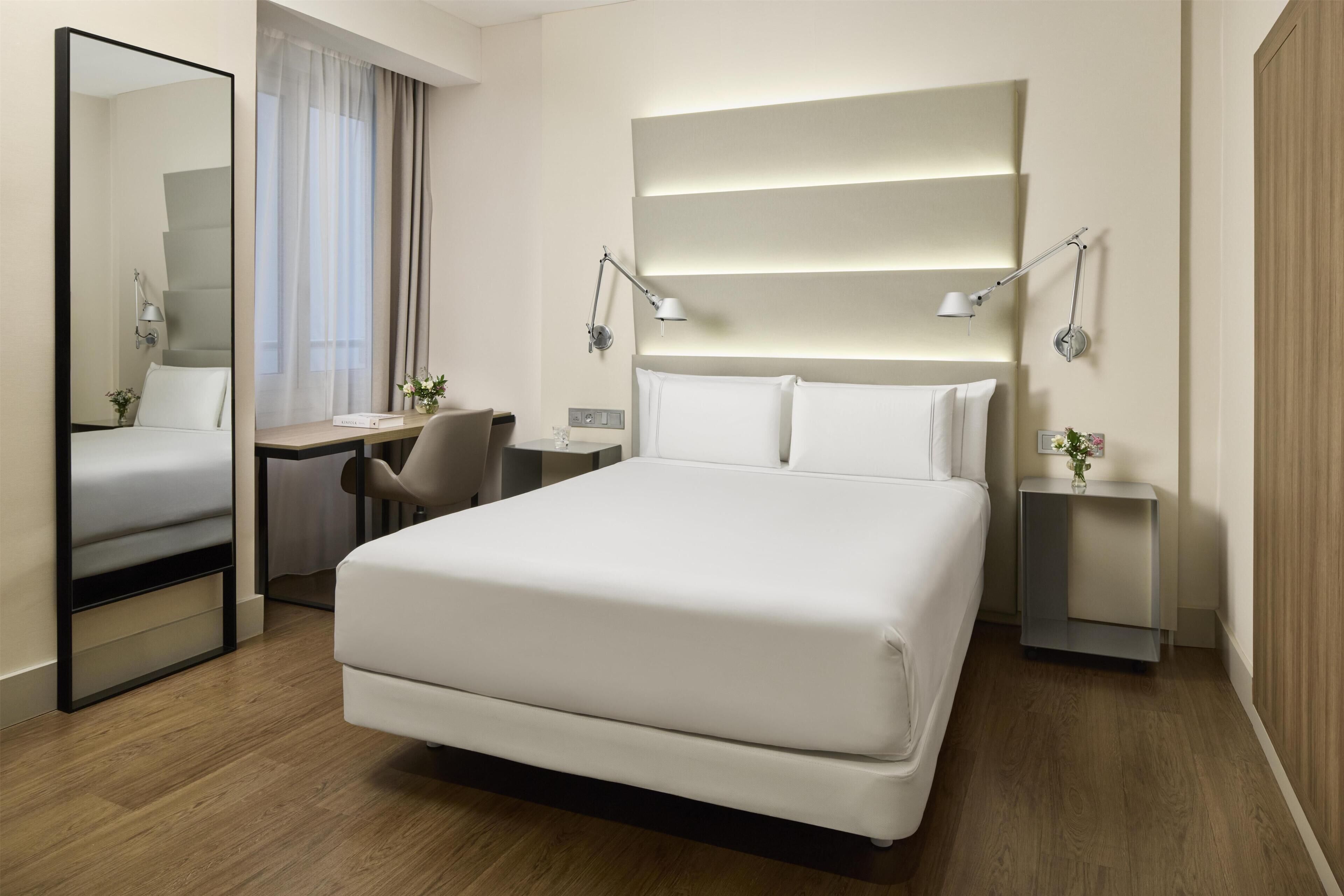 Habitación de hotel con una cama grande, mesitas de noche, un escritorio, una silla, un espejo y una ventana con cortinas.