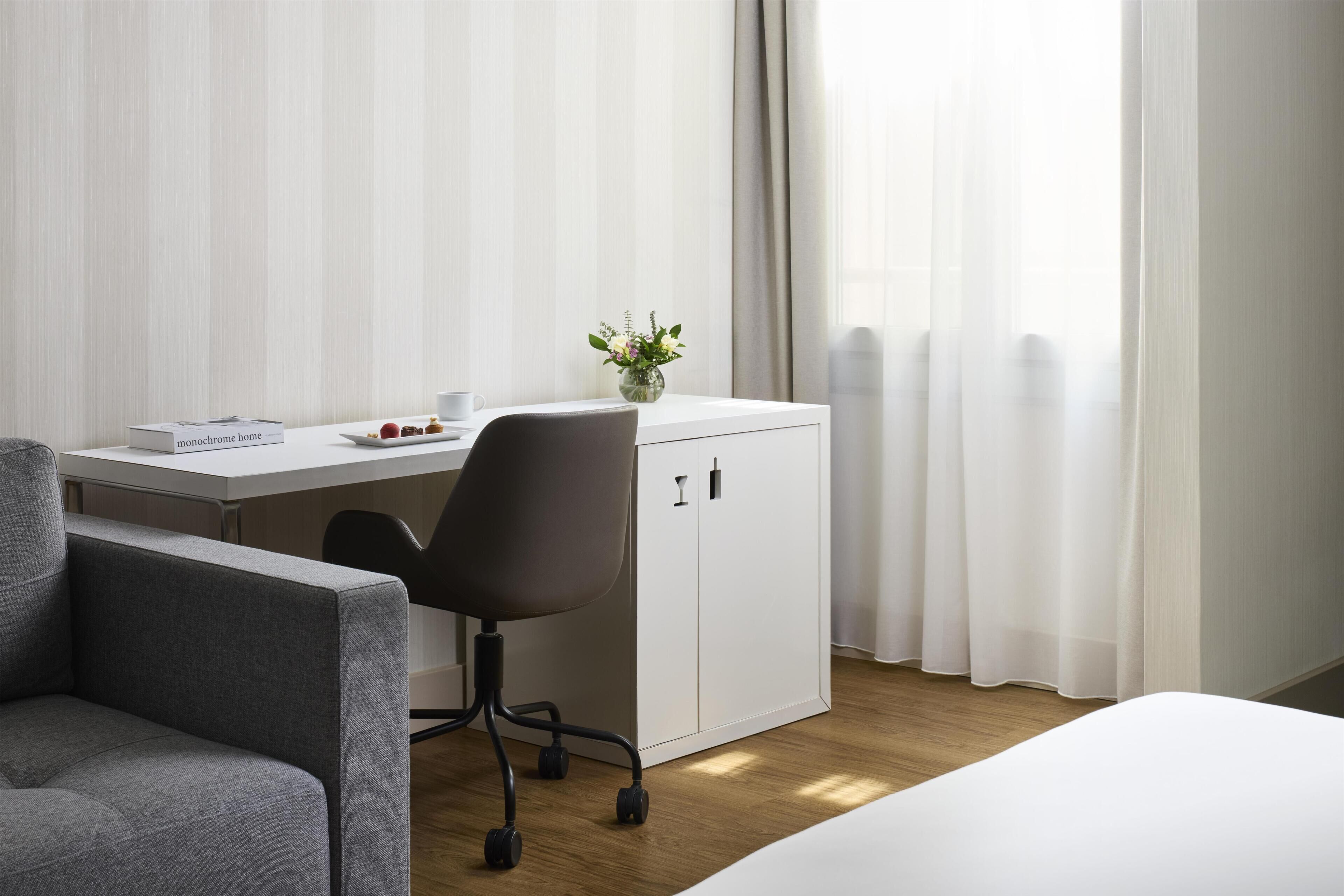 A hotel room with a bed, a desk, a chair, a small cabinet, and a window with curtains.