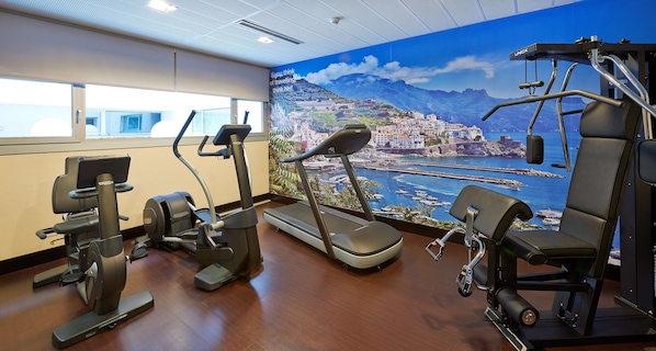 Une salle de sport équipée de machines de cardio, un mur décoré d’une fresque représentant une ville côtière, et une fenêtre offrant une vue panoramique.