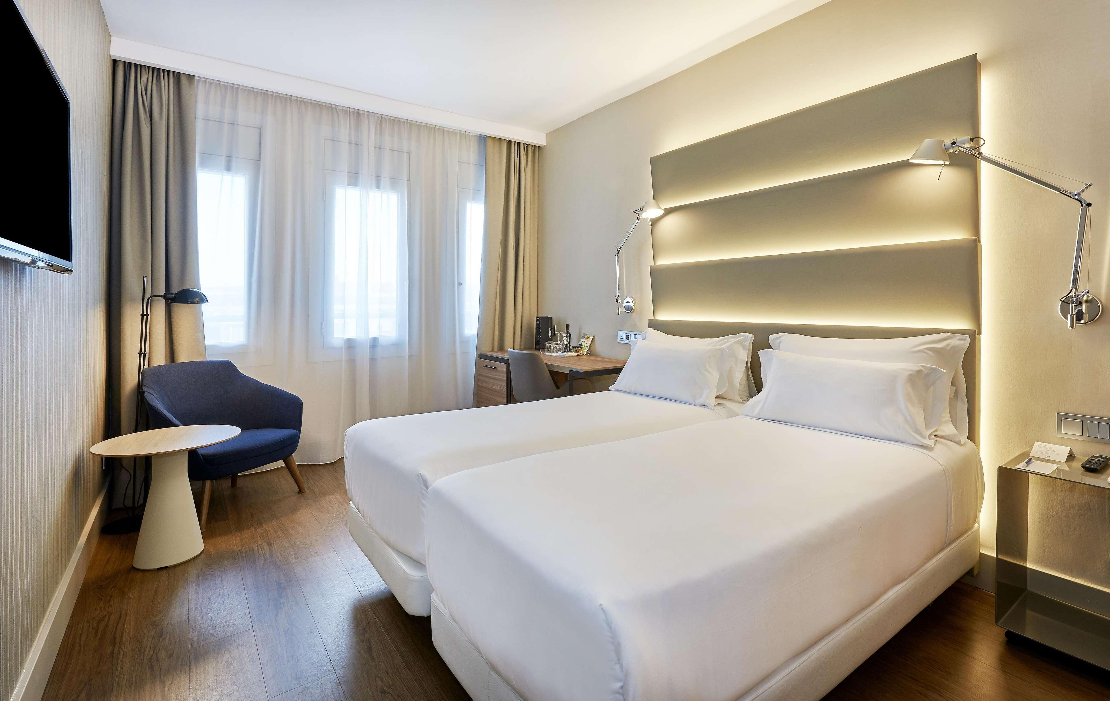 A hotel room with two beds, a desk, a chair, a TV, and a bedside table with a lamp.