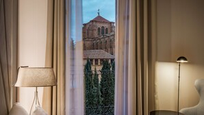 Habitación de hotel con una cama grande, una lámpara de noche, una mesita redonda y vistas a un edificio histórico a través de cortinas traslúcidas.