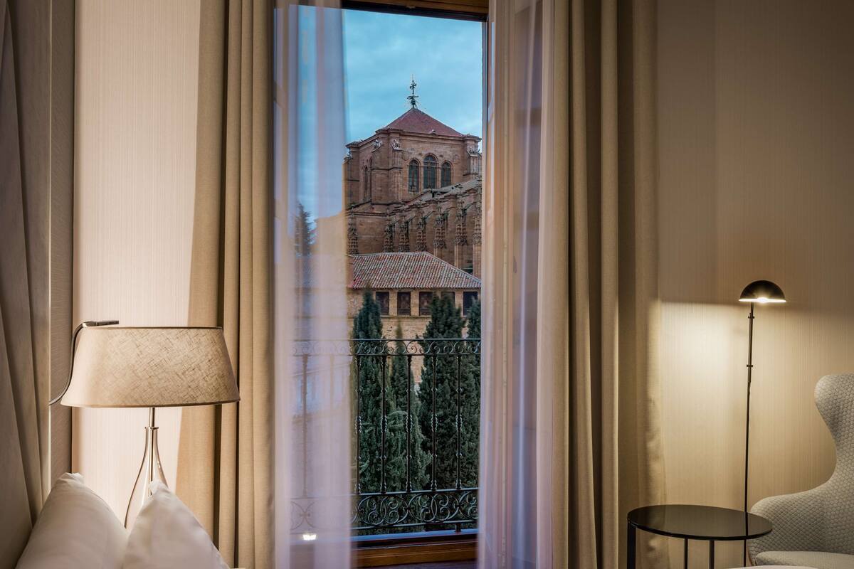 Une chambre d’hôtel avec un grand lit, une lampe de chevet, une petite table ronde et une vue sur un bâtiment historique, visible à travers des rideaux légers.