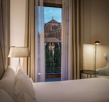 Habitación de hotel con una cama grande, una lámpara de noche, una mesita redonda y vistas a un edificio histórico a través de cortinas traslúcidas.