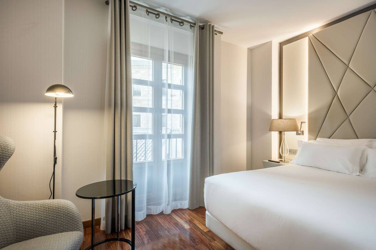 Une chambre d’hôtel avec un lit, une chaise, une petite table, une lampe et une fenêtre avec des rideaux.