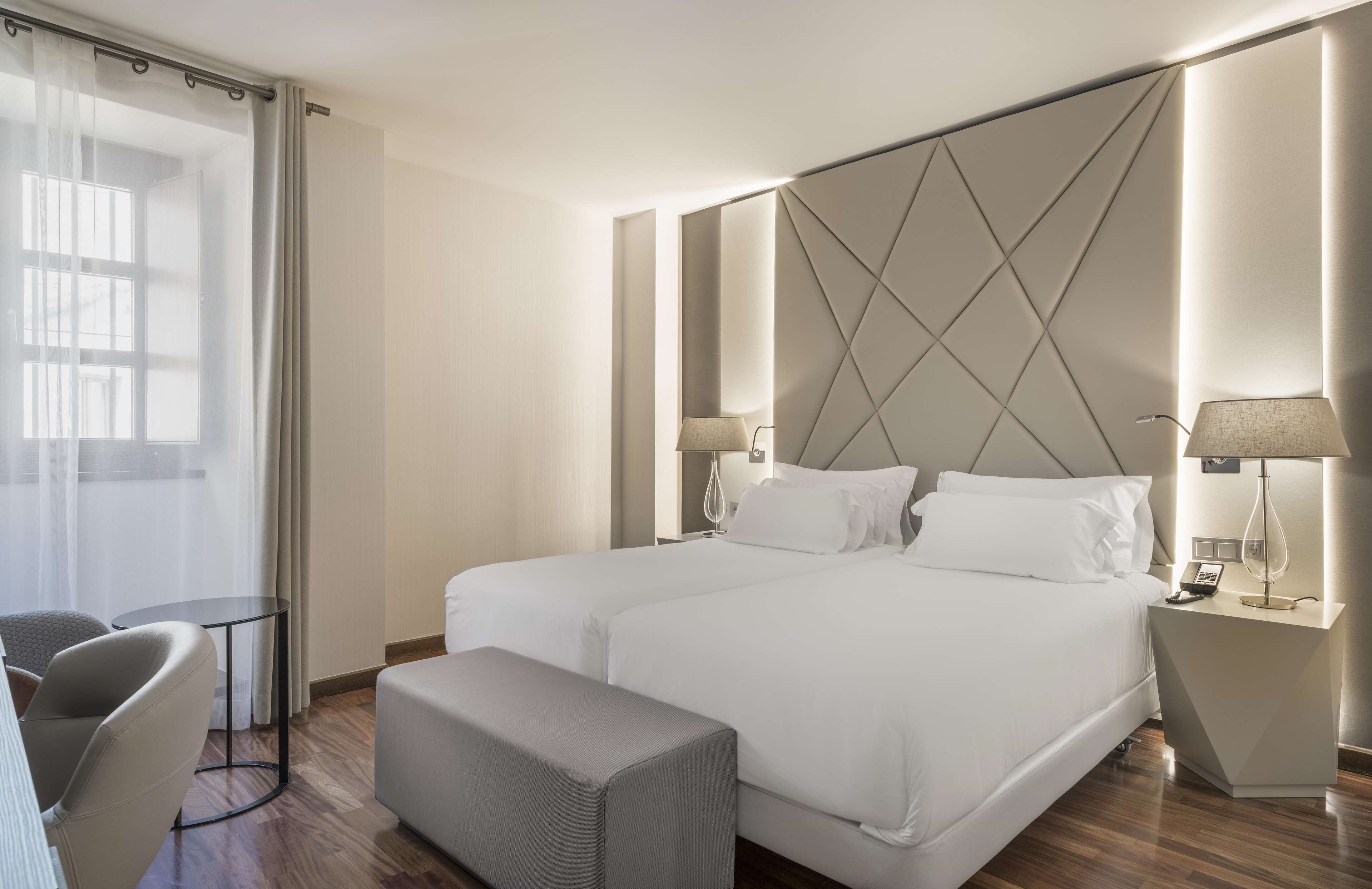 Une chambre d’hôtel moderne avec un grand lit, deux tables de chevet avec des lampes, une petite table ronde et un banc.