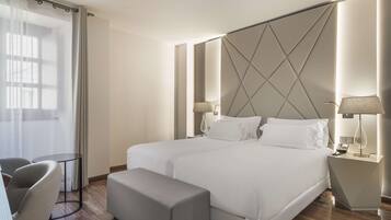 A modern hotel room with a large bed, two bedside tables with lamps, a small round table, and a bench.