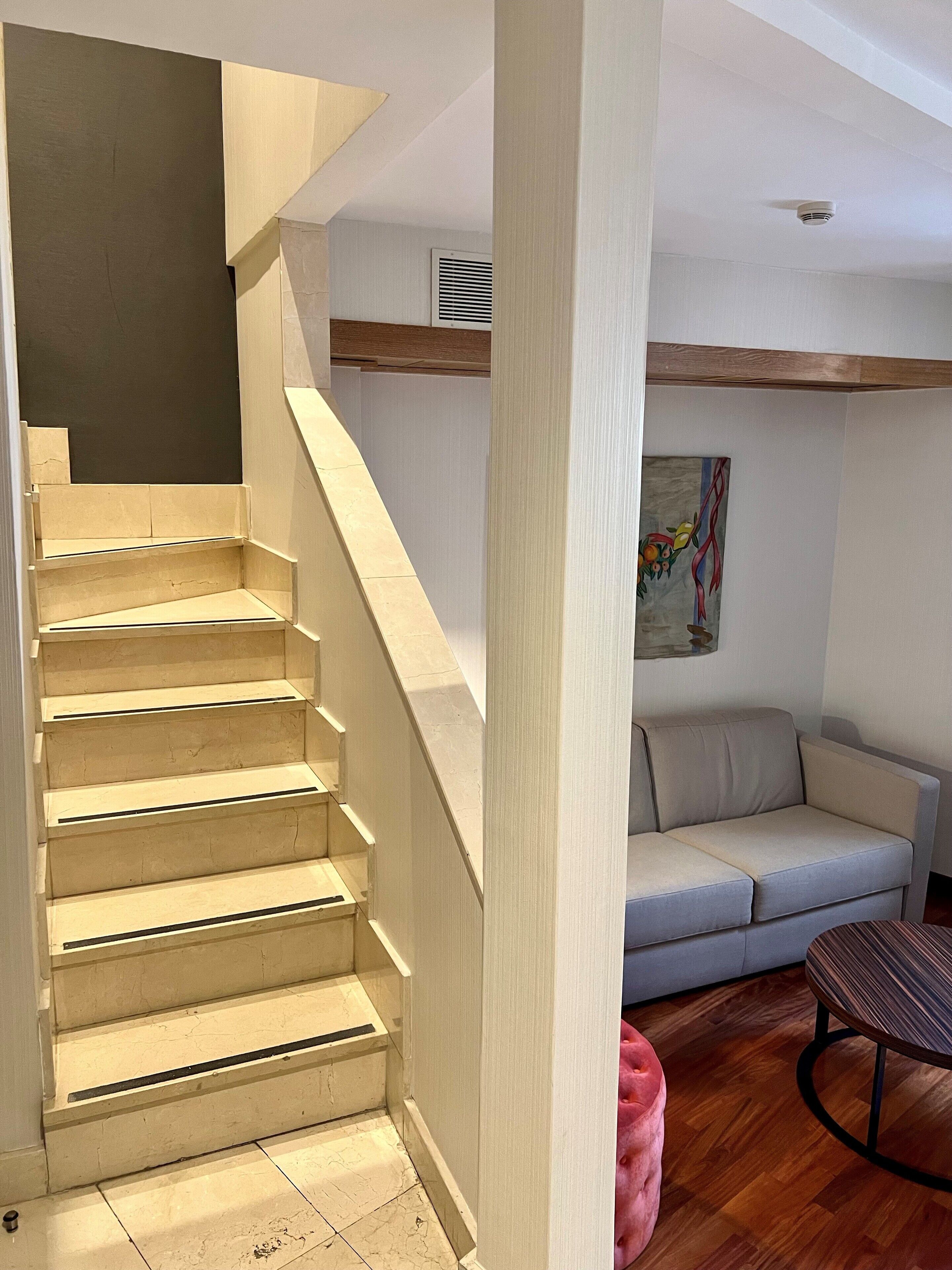 Eine Treppe mit beiger Farbe, eine weiße Wand mit einem Gemälde, ein graues Sofa und ein runder Tisch mit Streifenmuster.