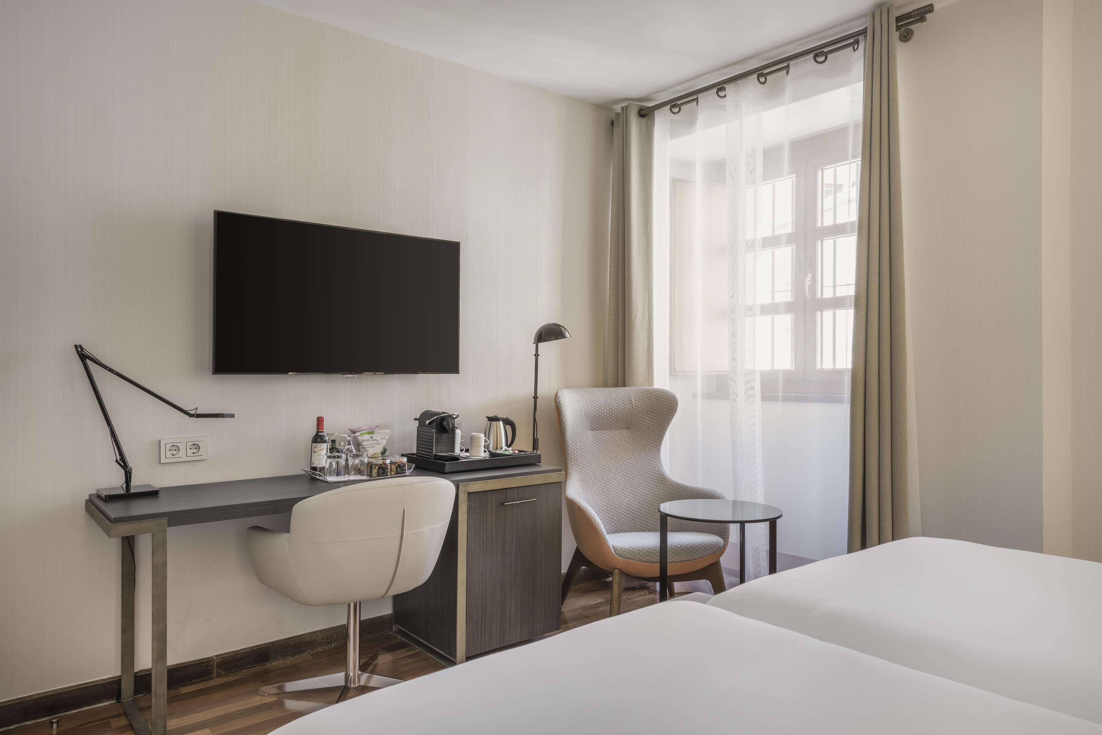 Habitación de hotel con cama, escritorio, silla, televisor, cafetera y ventana con cortinas.