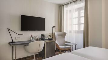 Habitación de hotel con cama, escritorio, silla, televisor, cafetera y ventana con cortinas.