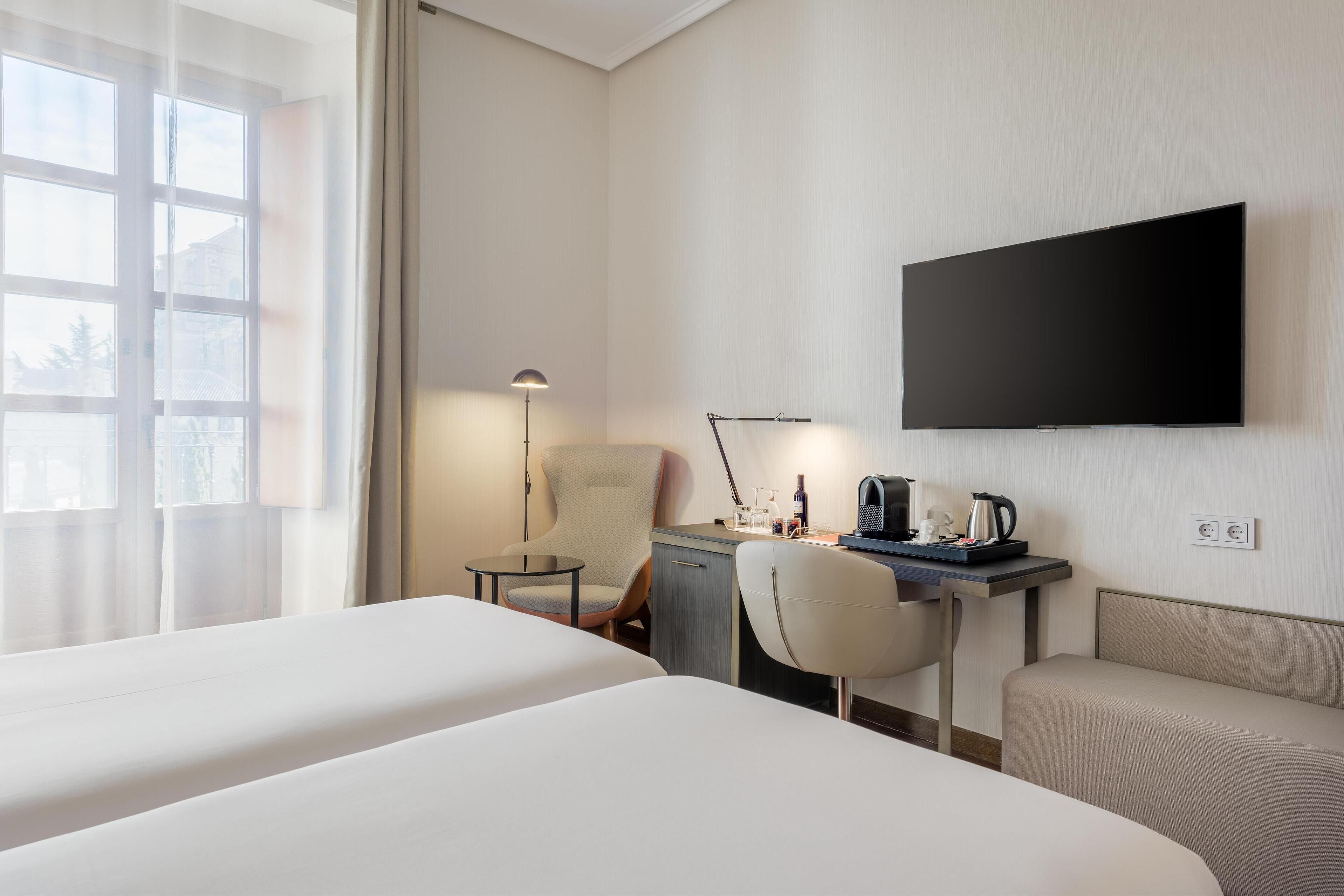A hotel room with two beds, a desk with a chair, a television, and a window with curtains.