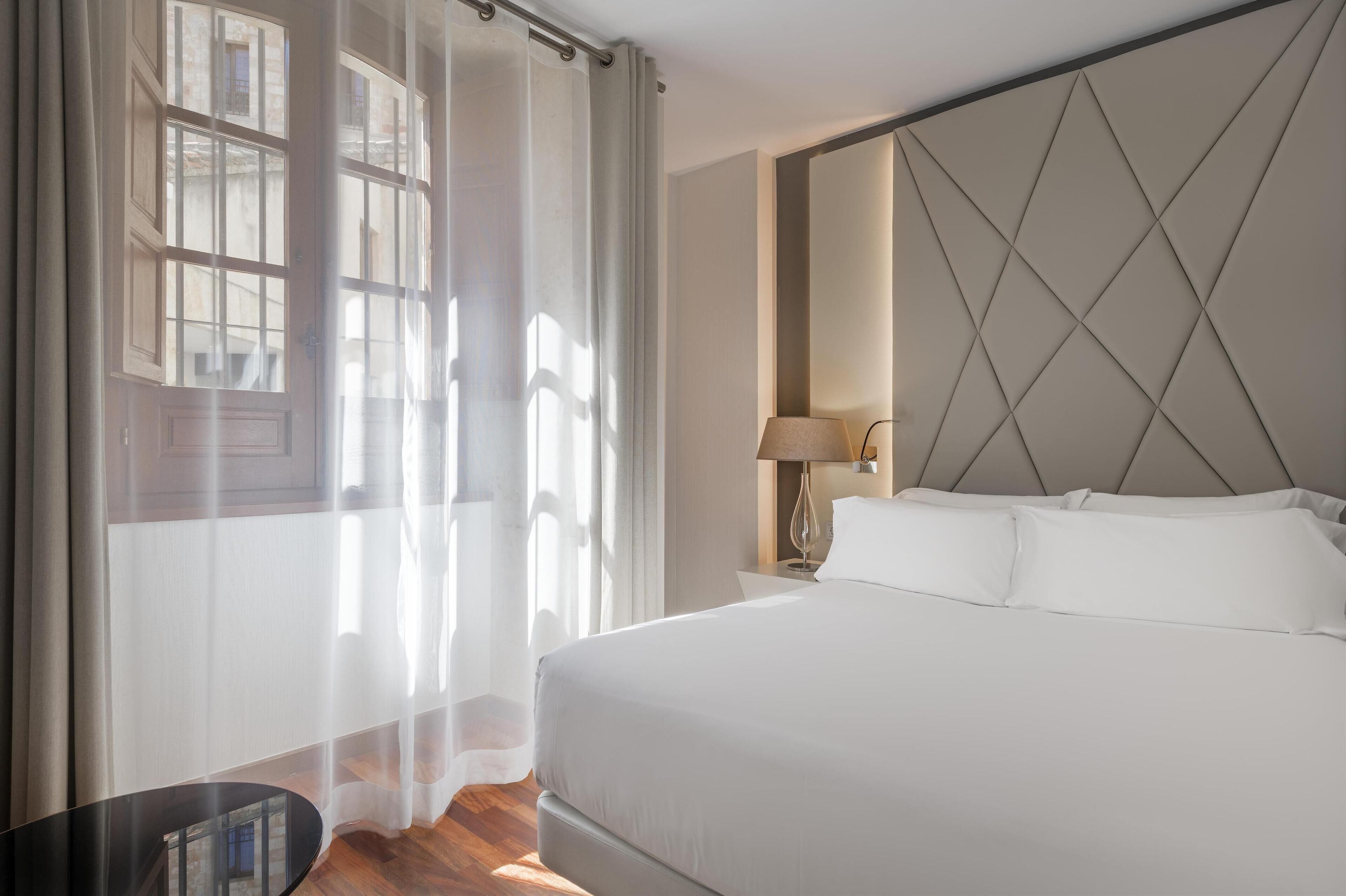 A hotel room with a large bed, a nightstand with a lamp, sheer curtains, and a view of the outside.