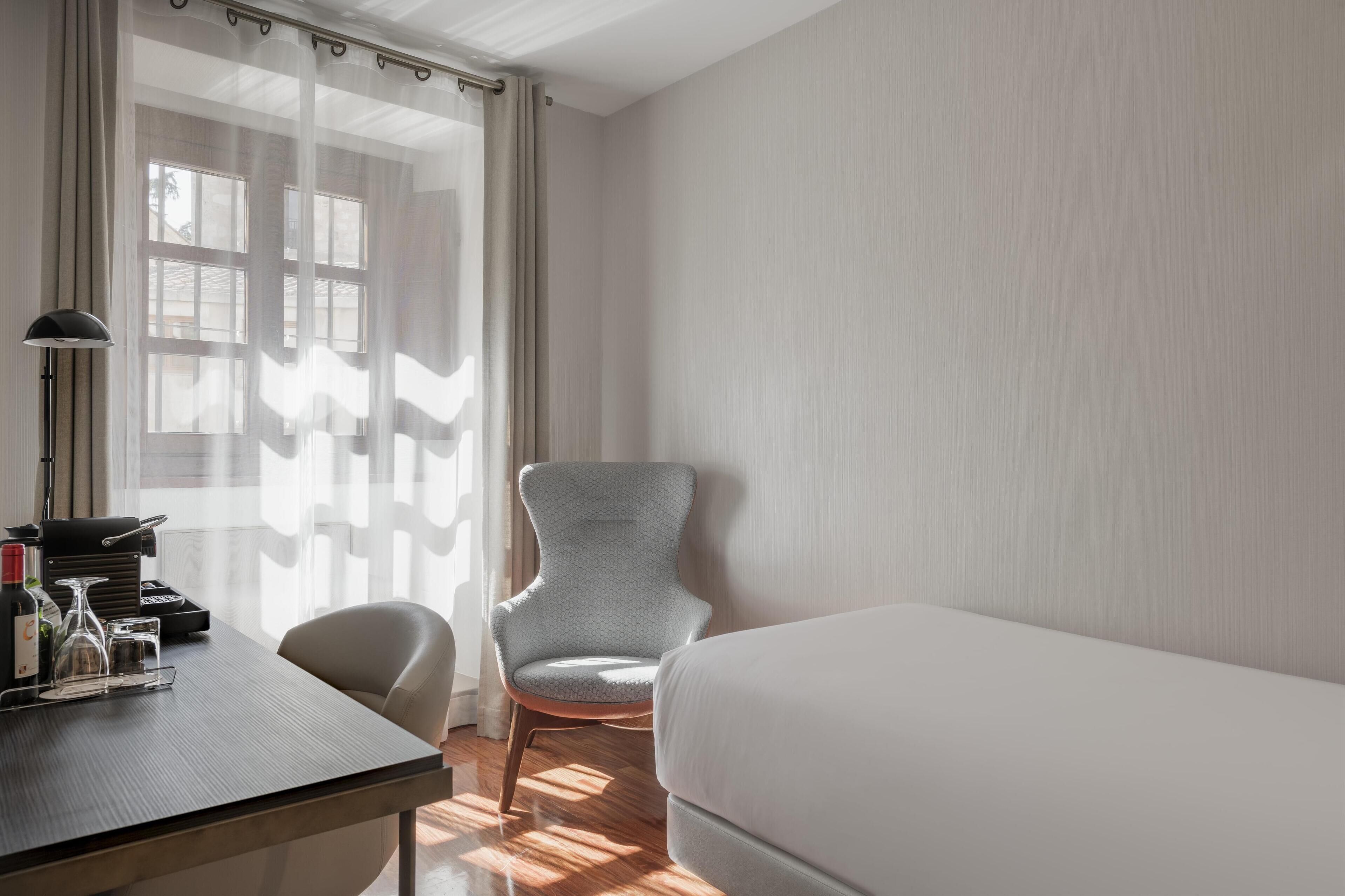 Una habitación de hotel con una cama, un escritorio con lámpara, una silla y una ventana con cortinas translúcidas.