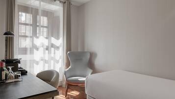 Una habitación de hotel con una cama, un escritorio con lámpara, una silla y una ventana con cortinas translúcidas.