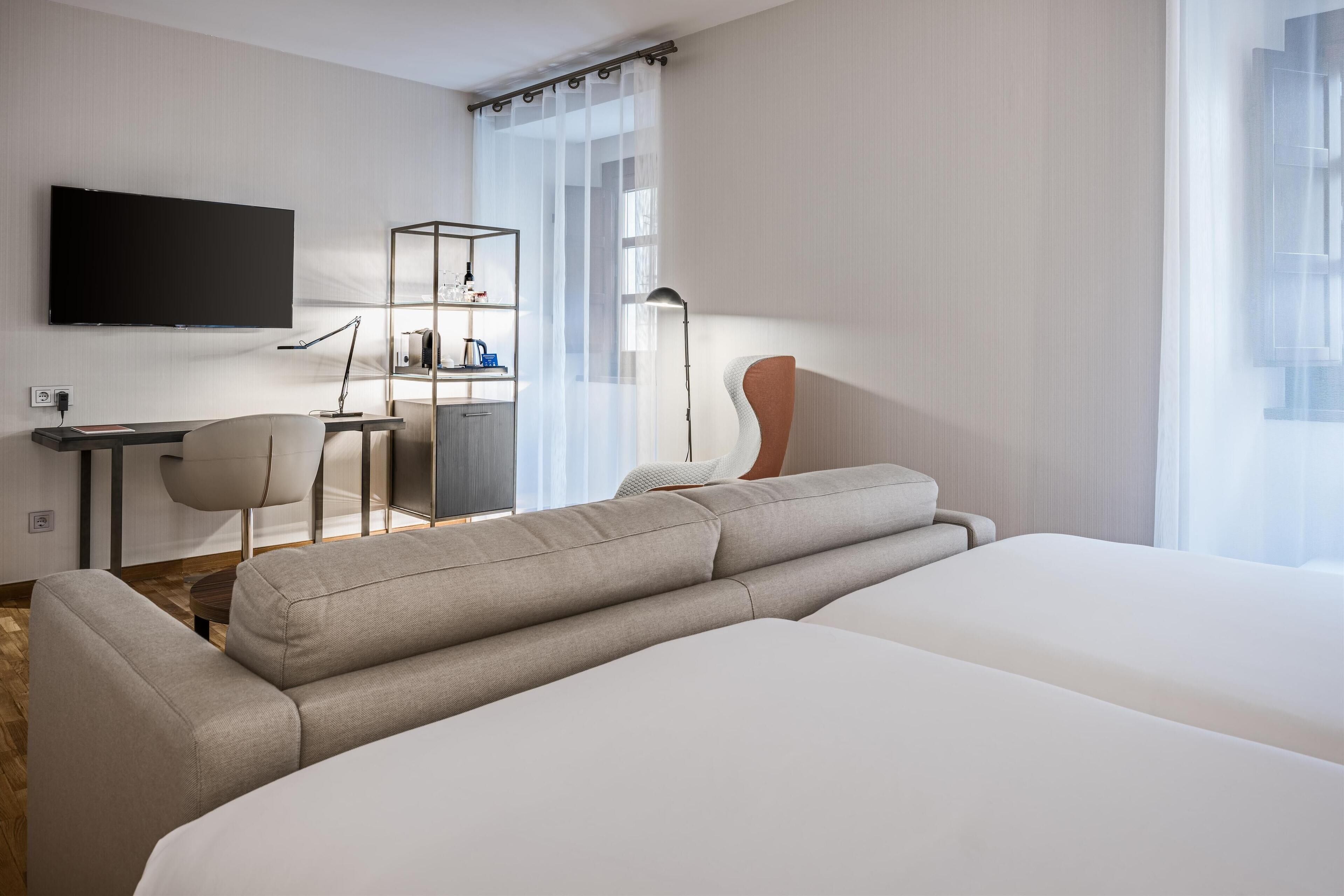 A modern hotel room with a large bed, a desk with a chair, a TV mounted on the wall, and a bookshelf.