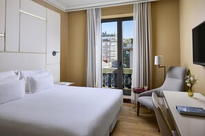 A hotel room with a large bed, a desk with a chair, a small table with a vase, and a view of buildings through the window.