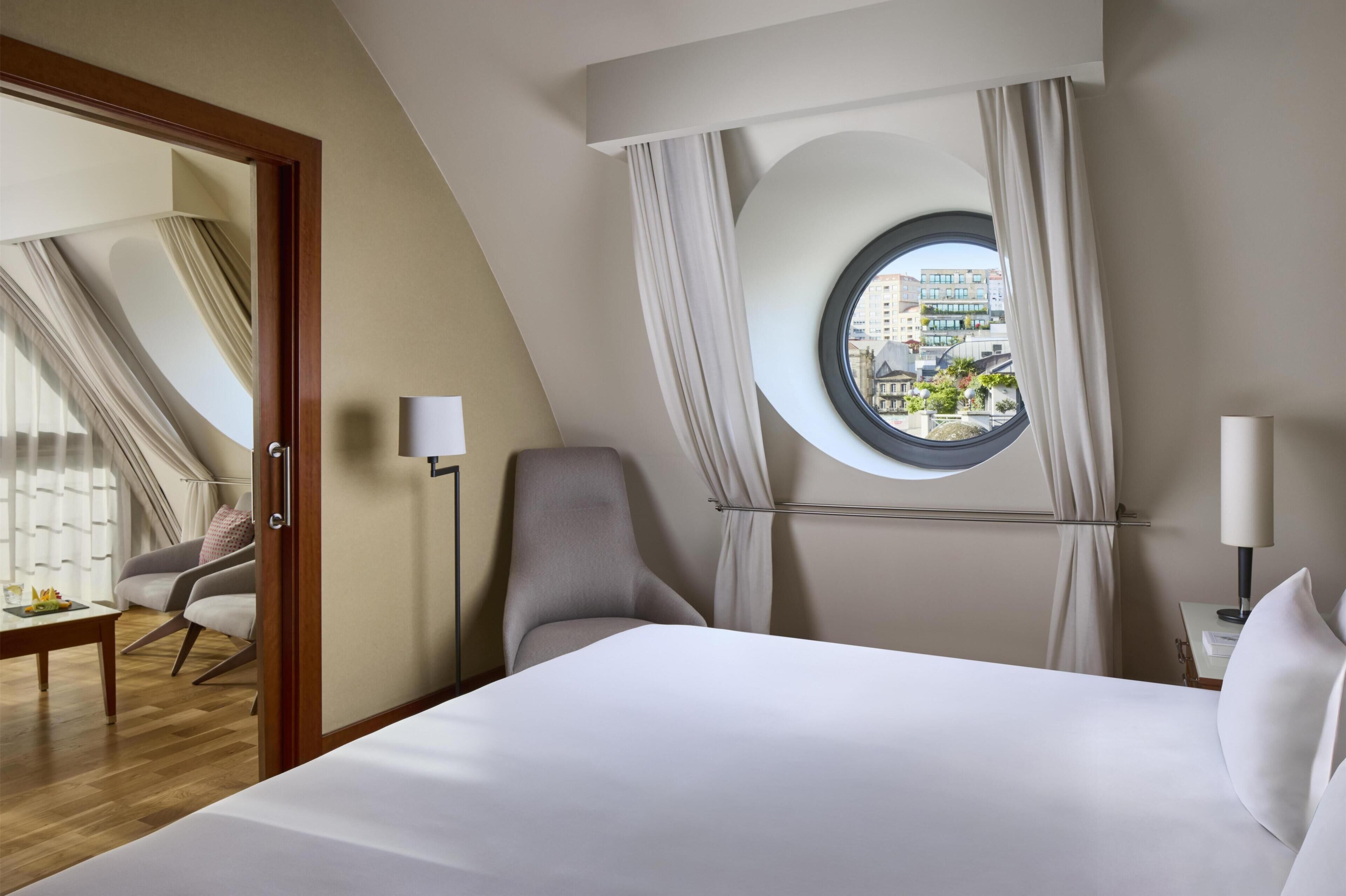 A hotel room with a bed, a chair, a round window with curtains, a table with a fruit plate, and a view of buildings outside.