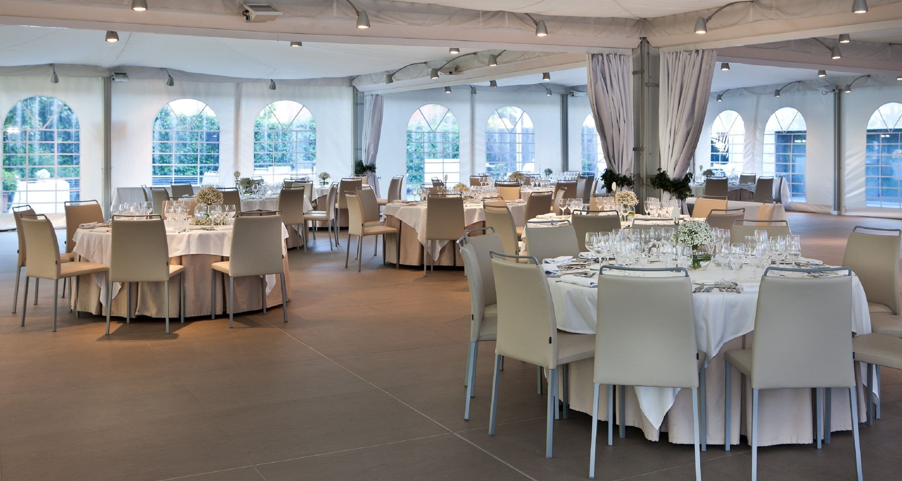 Een ruime balzaal met meerdere ronde tafels gedekt voor een diner, grote gewelfde ramen en een hoge plafond.