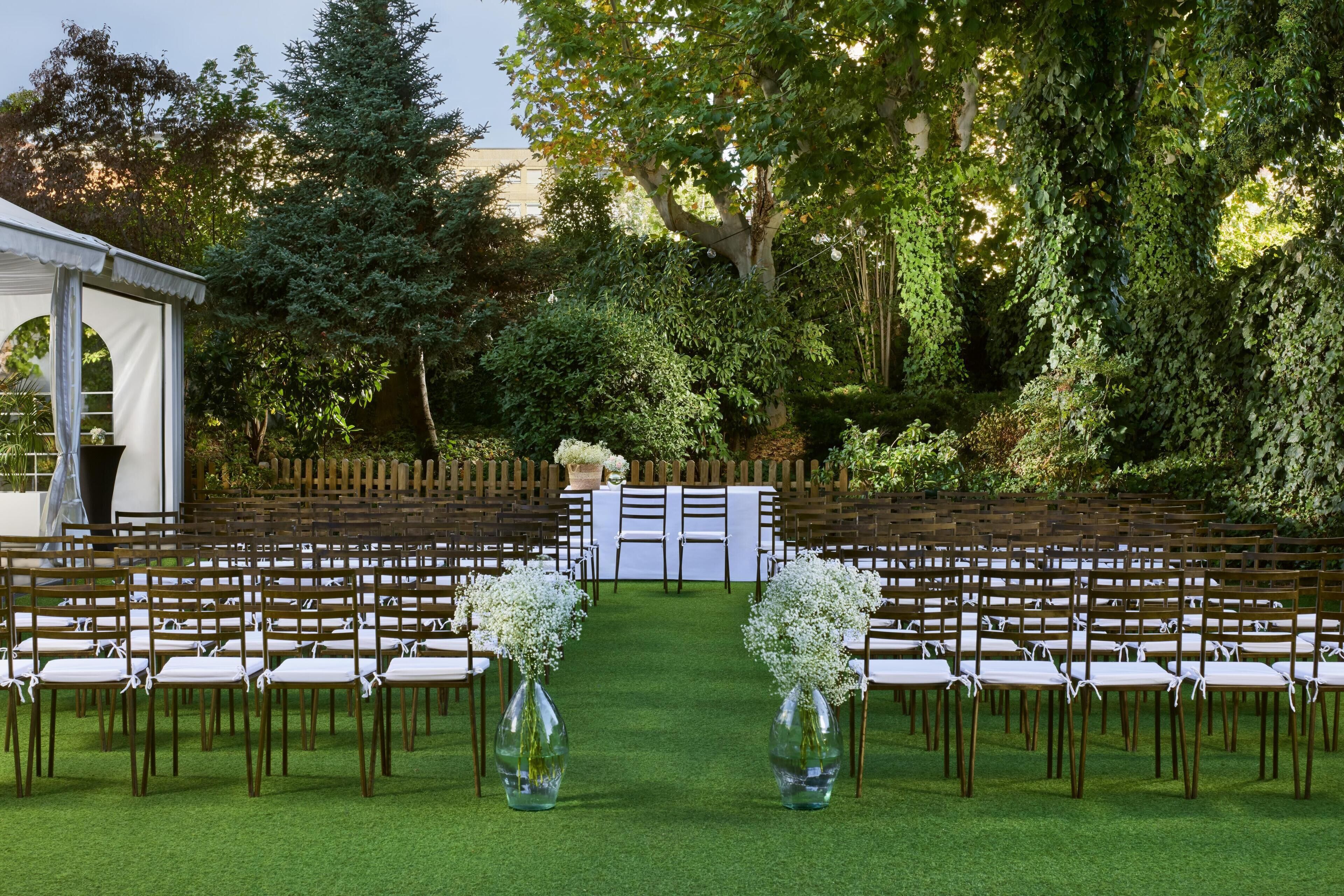 Un salón de bodas al aire libre con hileras de sillas de madera y manteles blancos, rodeado de exuberante vegetación y una carpa blanca.