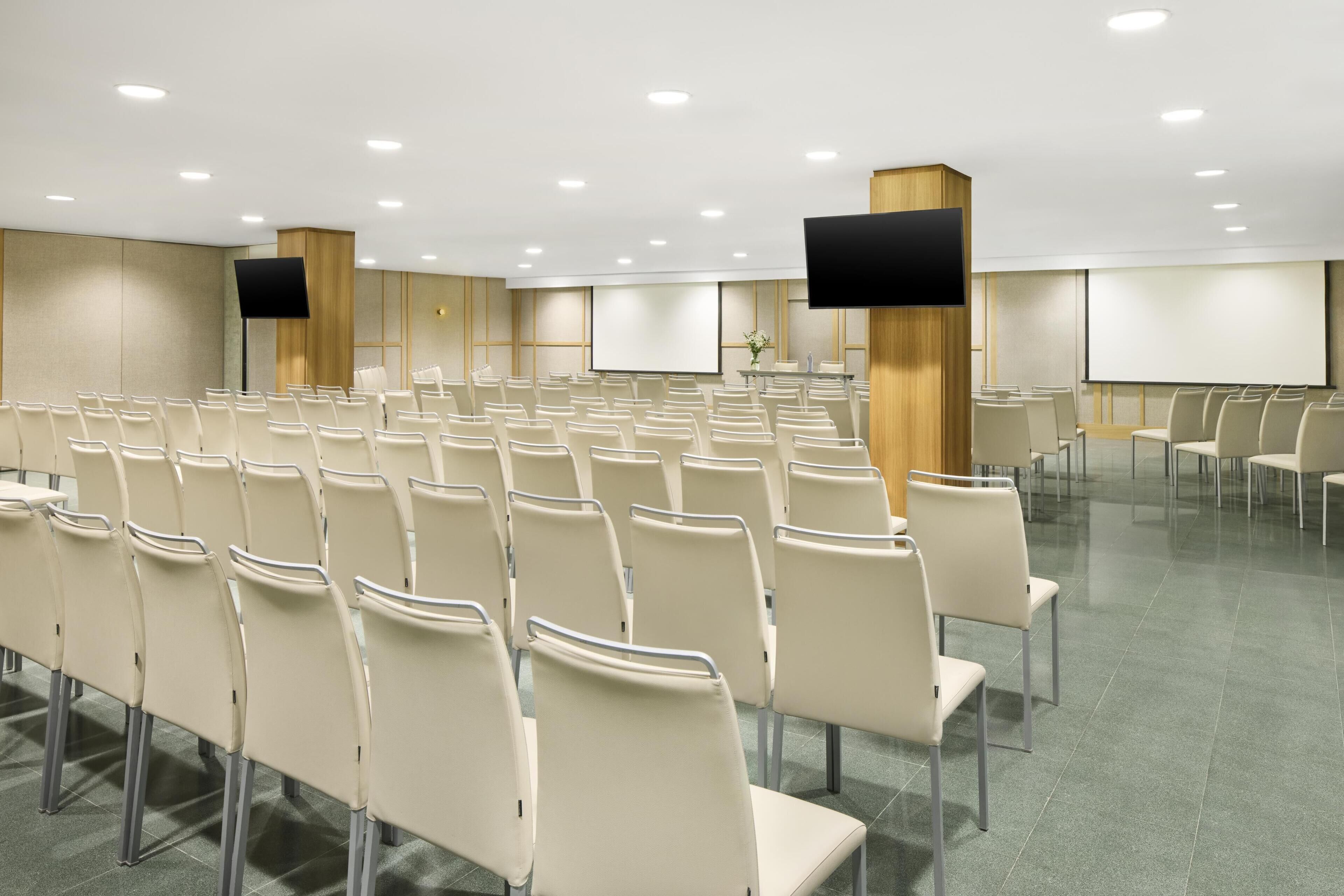Un salón de conferencias con filas de sillas, una pantalla grande y un proyector.
