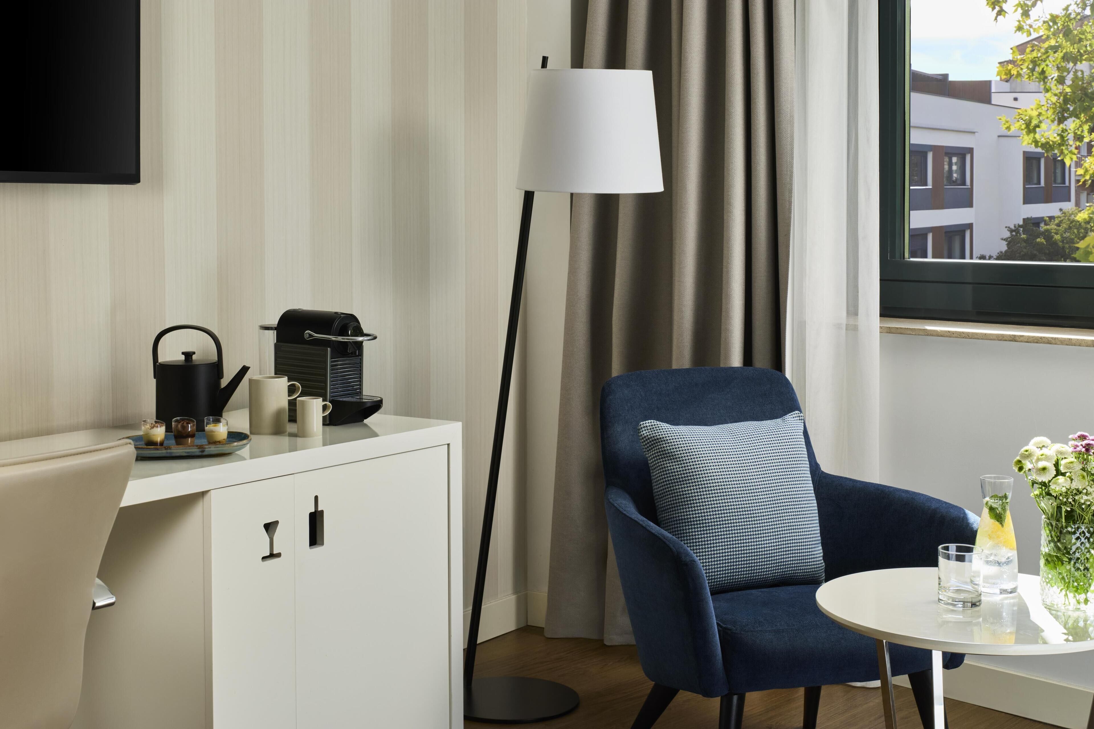 A modern hotel room with a sofa, a small table with a vase and glasses, a coffee machine, and a floor lamp.