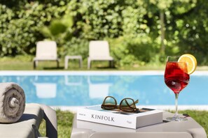 Escena junto a la piscina con un libro titulado 'KINFOLK' y un vaso de bebida roja con una rodaja de limón.