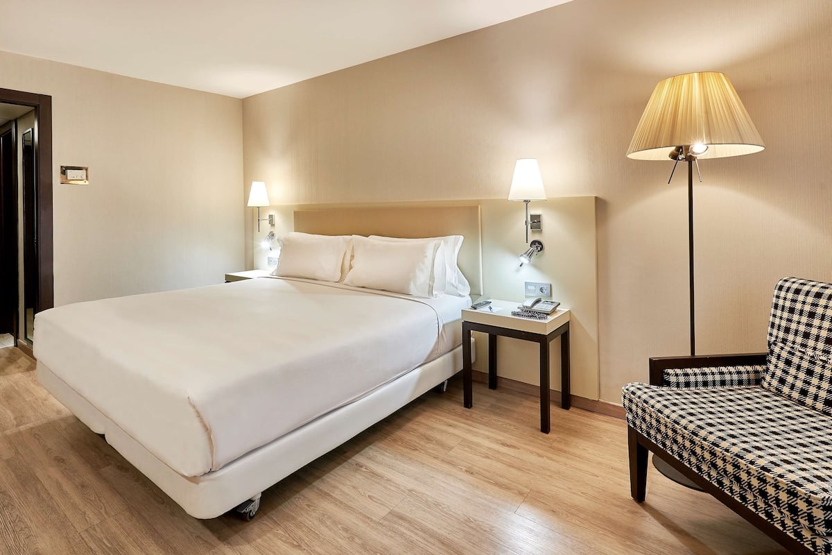 A hotel room with a large bed, bedside lamps, a small table with a phone and remote, and a patterned armchair.