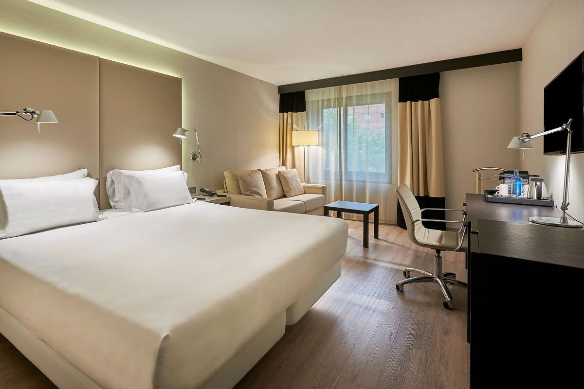 A hotel room with a large bed, a desk, a chair, a small table, and a sofa.