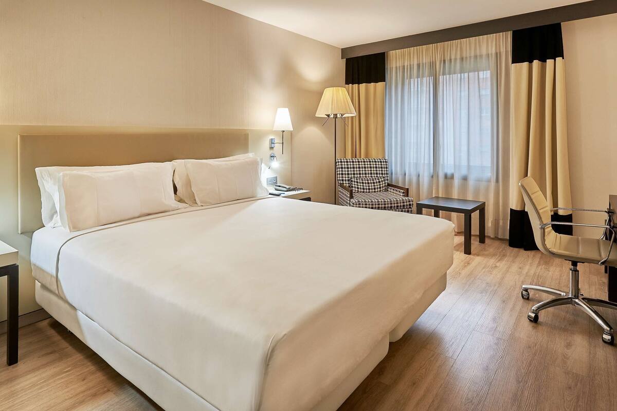 A hotel room with a large bed, two bedside tables, a desk with a chair, and a lamp.