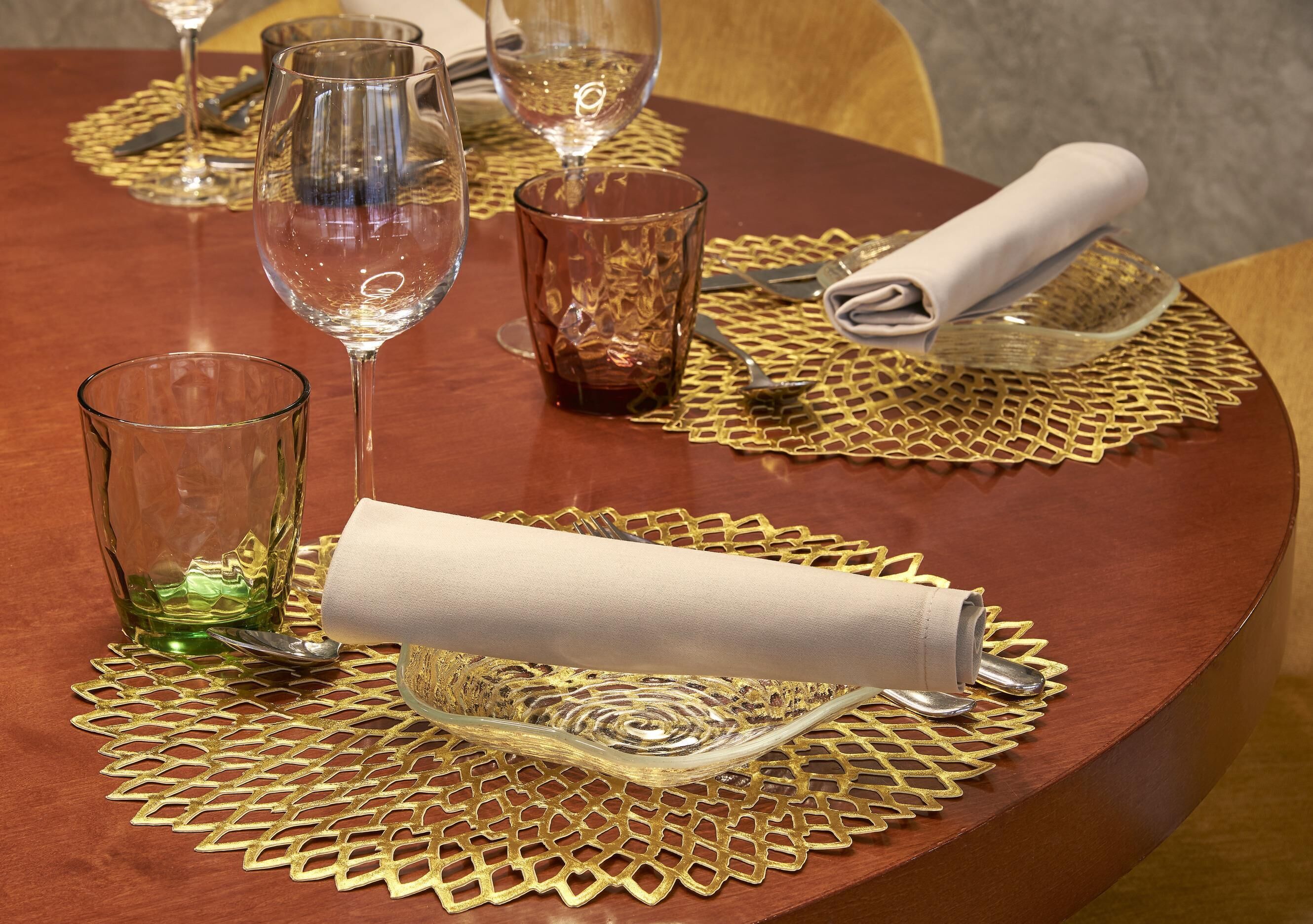A round dining table with a gold-patterned placemat, a folded white napkin, and a clear glass bowl.