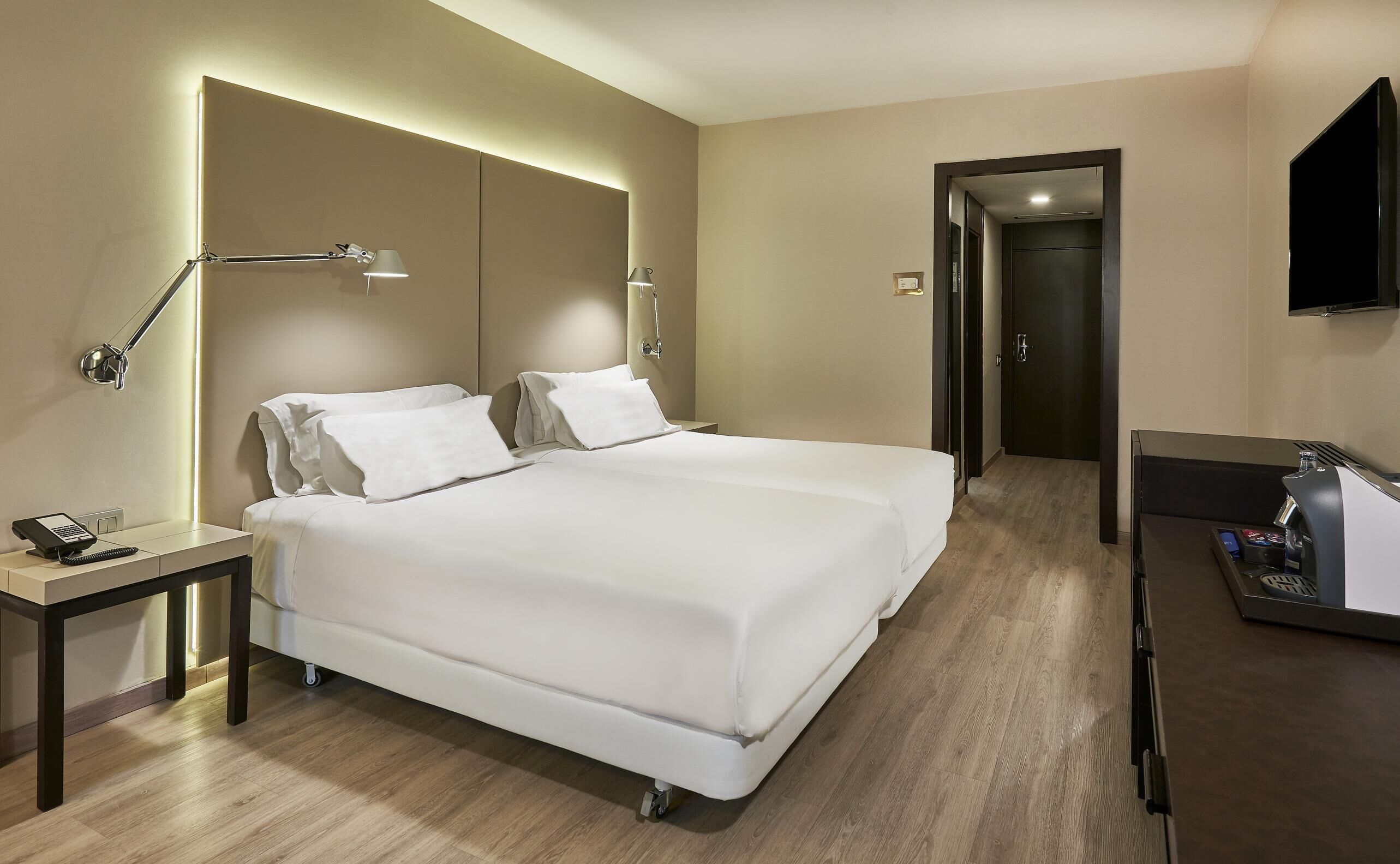 A hotel room with a large bed, bedside tables, a desk with a chair, a flat-screen TV, and a door leading to another room.