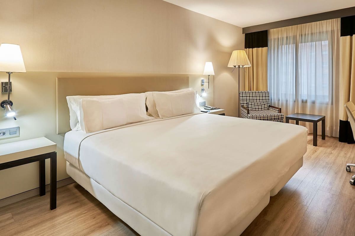 A hotel room with a large bed, bedside lamps, a desk, a chair, and a view of the outdoors through the window.