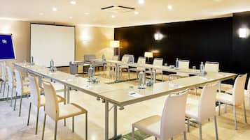 Un salón de conferencias con una larga mesa, sillas blancas, una pantalla de proyector y una pared decorativa negra.